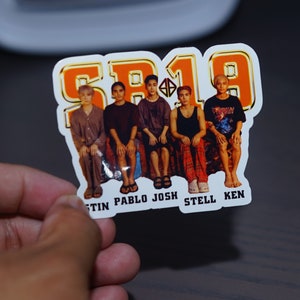 May include: A sticker with a group of five men in casual clothing. The sticker has an orange background with the text "SB19" in gold lettering. The names of the men are listed below the image: "Justin, Pablo, Josh, Stell, Ken."