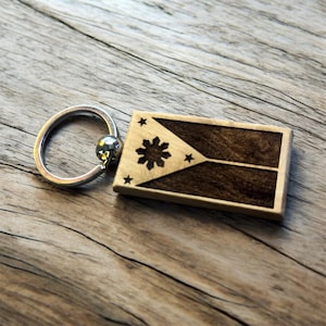 May include: Wooden keychain featuring the flag of the Philippines. The rectangular tag has a dark brown and light brown design with a silver key ring. The flag design includes a sun and three stars.