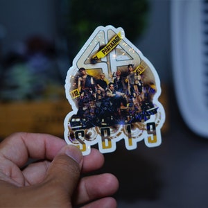 May include: A white sticker with a holographic finish featuring a group of people in front of a large number "44" with the text "SB19" below. The sticker has a yellow caution tape design with the text "PASILAN" on it.