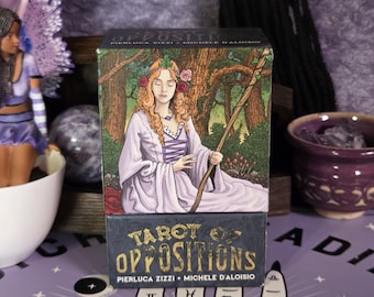 Pode incluir: Um baralho de cartas de tarô intitulado "Tarot of Oppositions" com uma ilustração de uma mulher com flores no cabelo. A caixa é escura com letras douradas. O baralho está sobre uma superfície roxa com um design de quiromancia.