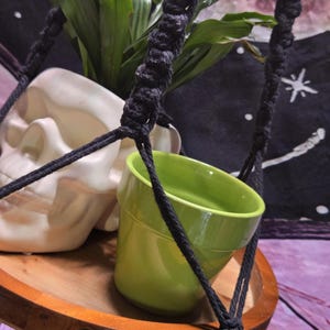 May include: A hanging macrame plant holder with a wooden tray base. It features a white skull-shaped planter, a green pot, and a plant with green leaves. The macrame is black, and the tray is a light brown color.