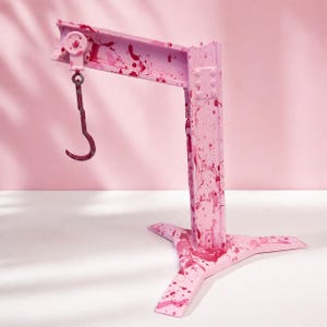 May include: A pink metal crane with a curved hook, splattered with red paint. The crane has a three-legged base and a horizontal beam with a pulley. The background is a solid pink color.
