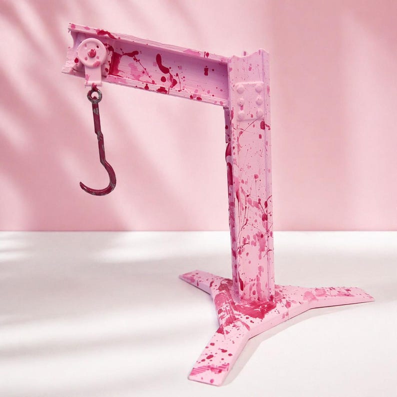 May include: A pink metal crane with a curved hook, splattered with red paint. The crane has a three-legged base and a horizontal beam with a pulley. The background is a solid pink color.