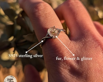 May include: A silver ring with a clear resin setting containing a dried flower, fur, and glitter. The ring is on a finger and the text "S925 sterling silver" and "fur, flower & glitter" is visible.