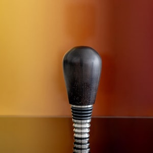 May include: A dark brown wooden bottle stopper with a rounded top and a silver metal base. The stopper has a textured appearance and is designed to fit into a bottle. The background is a gradient of orange and red.