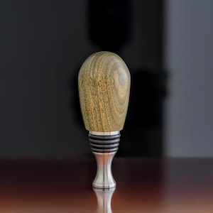 May include: A wooden bottle stopper with a green and brown wood grain pattern. The stopper has a silver metal base.