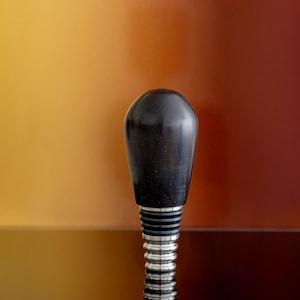 May include: A black wooden bottle stopper with a silver threaded base. The stopper is on a dark surface with a blurred background of orange, red, and yellow.