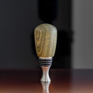 May include: A wooden bottle stopper with a green and brown wood grain pattern. The stopper has a silver metal base and a black rubber ring.