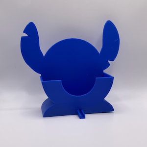 May include: A bright blue plastic desk organizer featuring a silhouette of a character with large, pointed ears and a round head. The holder has a curved front pocket for storing small items and a small, rectangular tab extending from the base.