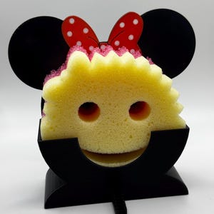 May include: A yellow sponge with two eye holes and a smiling mouth, held in a black holder. The sponge has a red bow with white polka dots on top. The holder has two black Mickey Mouse ears.