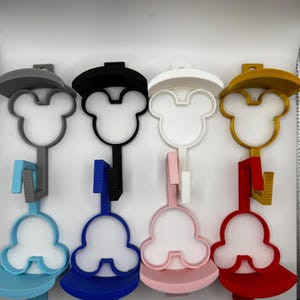 Puede incluir: Una colección de coloridos soportes para cepillos de dientes con forma de Mickey Mouse. Cada soporte presenta un diseño de sombrero y una base a juego, con colores como gris, negro, blanco, dorado, azul, rosa y rojo. Los soportes están diseñados para sujetar un cepillo de dientes.