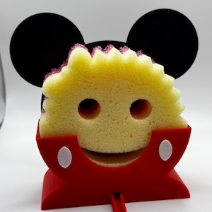 Puede incluir: Una esponja amarilla con un diseño de cara sonriente, sujeta en un soporte rojo con detalles blancos. El soporte tiene un diseño inspirado en Mickey Mouse con orejas circulares negras. La esponja tiene un borde rosa.