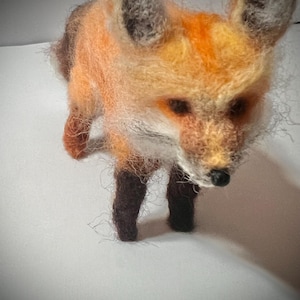 May include: A needle felted fox figurine with orange fur, black legs, and a white chest. The fox is standing on a white surface.