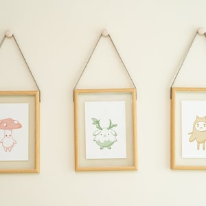 May include: Three framed illustrations of cartoon characters. The first illustration is of a pink mushroom with a face, the second is of a green creature with antlers, and the third is of a brown creature with a hood.