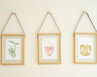 Puede incluir: Tres ilustraciones enmarcadas de animales de dibujos animados colgadas en una pared de color beige claro. Las ilustraciones están en un marco de madera clara con una correa de cuero marrón. La primera ilustración es un gato de dibujos animados verde con una planta que le crece de la cabeza. La segunda ilustración es un gato de dibujos animados rosa con una planta que le crece de la cabeza. La tercera ilustración es un gato de dibujos animados amarillo con una planta que le crece de la cabeza.