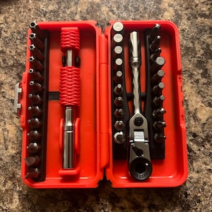 May include: An open red toolbox containing a variety of screwdriver bits and a ratchet. The bits are arranged in rows, and the ratchet is silver and black. A red handle is also included.