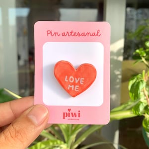 May include: A handmade ceramic pin in the shape of a red heart with the words "LOVE ME" in white. The pin is displayed on a pink card that reads "Pin artesanal" and features the "piwi cerámicas" logo.