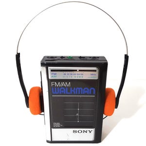 May include: A black and silver Sony FM/AM Walkman cassette player with a radio frequency display. The device is attached to a pair of black headphones featuring bright orange foam ear pads and a thin, adjustable metal headband.
