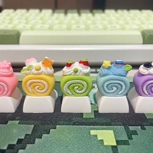 Puede incluir: Cinco teclas de color con temática de pastel con diferentes coberturas, incluyendo un pastel rosa con flores, un pastel amarillo con una estrella, un pastel verde con una fresa, un pastel azul con una estrella y un pastel morado con una estrella.