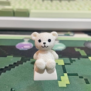 May include: A white teddy bear keycap with black eyes and a black nose sits on a white keycap base. The keycap is on a green and black mousepad.