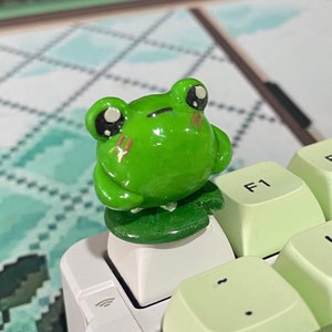 Puede incluir: Una figurita de rana verde con ojos grandes y una pequeña boca, sentada sobre una base de hoja verde. La rana está encima de un teclado verde claro con teclas blancas etiquetadas como F1 y F2.