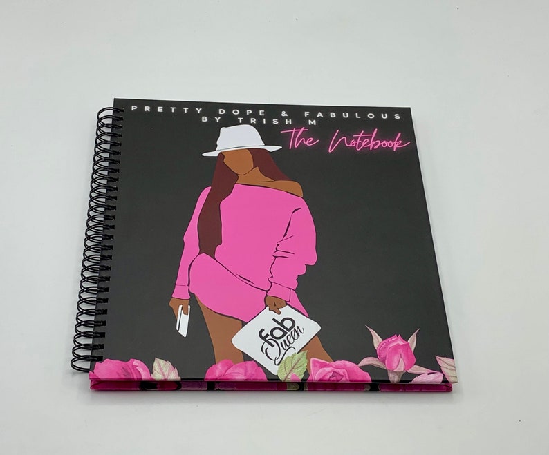 Peut inclure: Un carnet &agrave; spirales noir avec une illustration rose et noire d'une femme portant un chapeau blanc et une robe rose. Le carnet porte le texte "Pretty Dope & Fabulous by Trish M The Notebook" et "Fab Queen" sur la couverture.