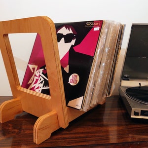 May include: A wooden record stand with a stack of vinyl records. The record stand is made of light wood and has a simple design. The records are stacked vertically and are visible through the open front of the stand. The record stand is sitting on a wooden surface.