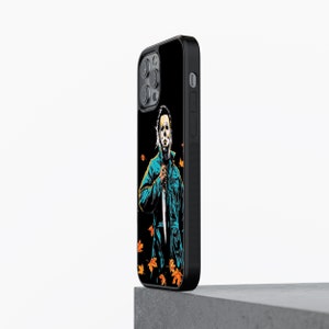 May include: Black phone case with a graphic of a masked figure holding a knife, surrounded by orange and yellow leaves. The figure is wearing a blue jumpsuit.