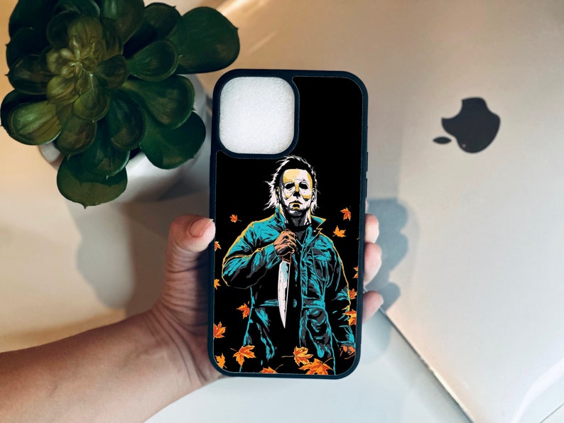 May include: Black phone case with a graphic of Michael Myers from the Halloween movies. He is holding a knife and standing in front of a background of orange and yellow leaves.