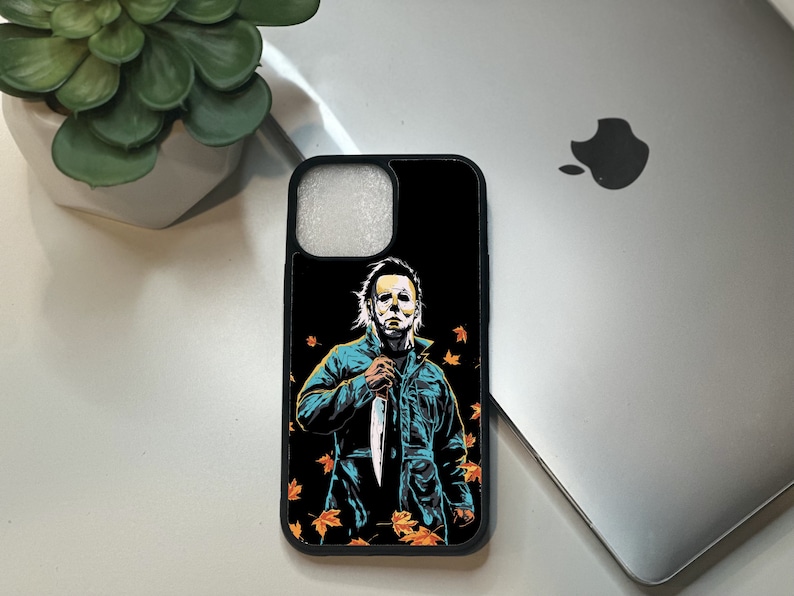 May include: Black phone case with a graphic of Michael Myers from the Halloween movies holding a knife. The background is black with orange and yellow leaves.