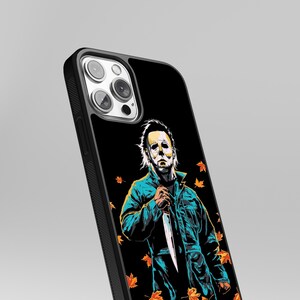 May include: Black iPhone case with a graphic of Michael Myers from the Halloween movies. The graphic is in a cartoon style and features Michael Myers holding a knife. The background is black with orange and yellow leaves.