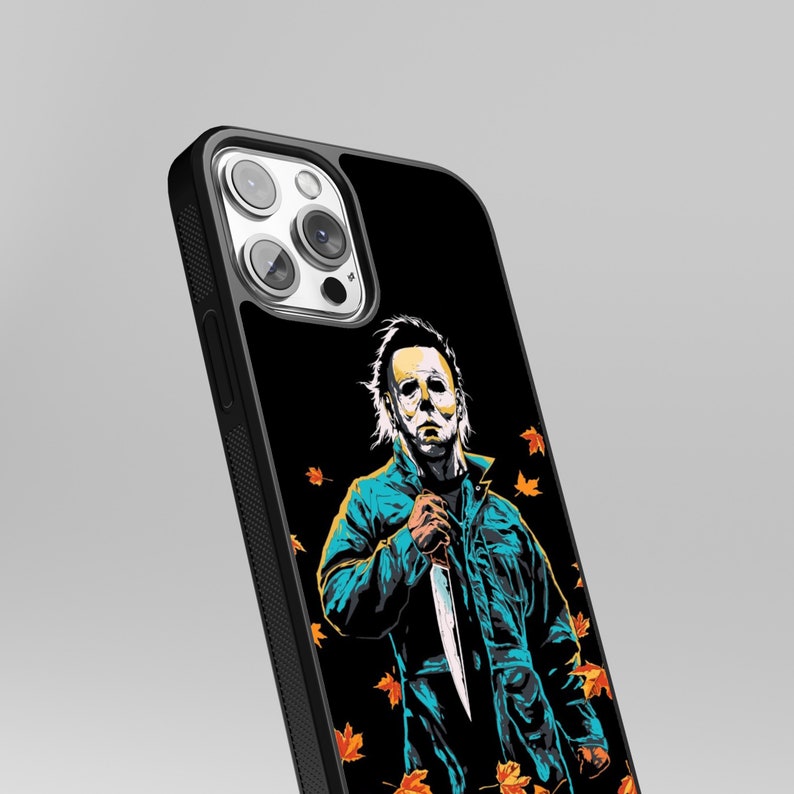 May include: Black iPhone case with a graphic of Michael Myers from the Halloween movies. The graphic is in a cartoon style and features Michael Myers holding a knife. The background is black with orange and yellow leaves.