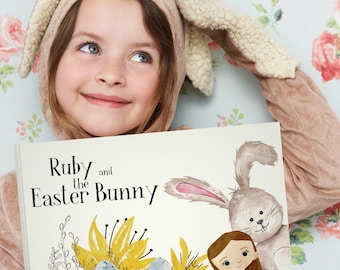 May include: A young girl wearing a brown bunny costume holds a book titled "Ruby and the Easter Bunny". The book features an illustration of a girl and a bunny rabbit.