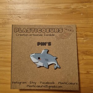 May include: A light blue and white cartoon shark pin with a smiling face. The pin is on a brown card with the text "Plasticoeurs Creation artisanale familiale Pin's Instagram Etsy Facebook Plasticoeurs Plasticoeurs@gmail.com"