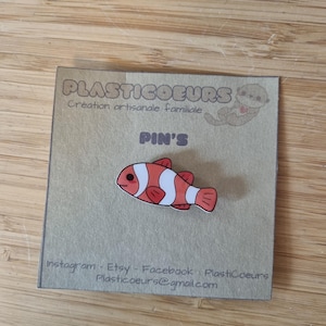 Puede incluir: Un pequeño pin de pez de dibujos animados naranja y blanco con ojos negros. El pin está en una tarjeta marrón con el texto "Plasticoeurs Creation artisanale familiale Pin's Instagram - Etsy - Facebook: Plasticoeurs Plasticoeurs@gmail.com"