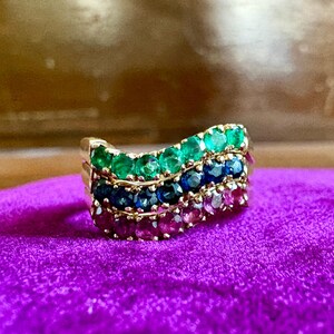 May include: A gold ring featuring three wavy, stacked rows of round-cut gemstones. The top row consists of green emeralds, the middle row contains deep blue sapphires, and the bottom row is set with vibrant pink rubies in a prong setting.