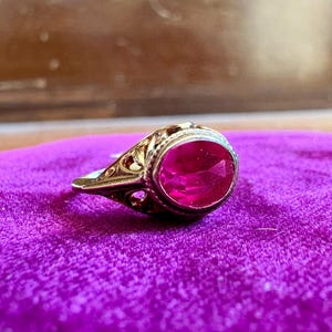 May include: A gold ring featuring an oval-cut, vibrant pinkish-red gemstone set in a bezel. The metal band displays intricate, openwork filigree detailing along the sides, extending from the central stone setting to the shank of the ring.