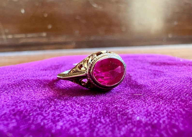 May include: A gold ring featuring an oval-cut, vibrant pinkish-red gemstone set in a bezel. The metal band displays intricate, openwork filigree detailing along the sides, extending from the central stone setting to the shank of the ring.