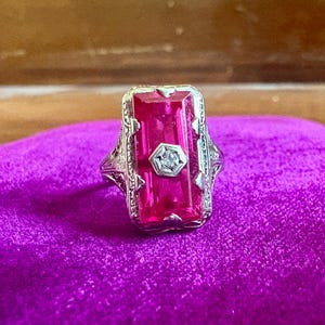 May include: A silver-toned ring featuring a large, rectangular, vibrant pink gemstone set vertically. A small, clear, round diamond is centered within a hexagonal frame on top of the pink stone. The metal band displays intricate, filigree-style metalwork.
