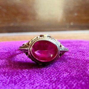 May include: A ring featuring an oval-shaped, vibrant pink gemstone set in a metallic band with intricate, decorative filigree metalwork along the sides. The setting is detailed with small, raised patterns that frame the central stone.