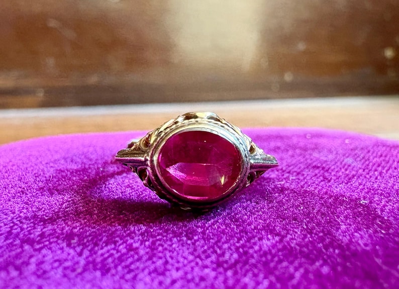 May include: A ring featuring an oval-shaped, vibrant pink gemstone set in a metallic band with intricate, decorative filigree metalwork along the sides. The setting is detailed with small, raised patterns that frame the central stone.