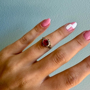May include: A gold ring featuring an oval-shaped red gemstone set in a decorative metal frame. The band has intricate filigree detailing along the sides, surrounding the central stone which is held in place by a simple metal bezel setting.