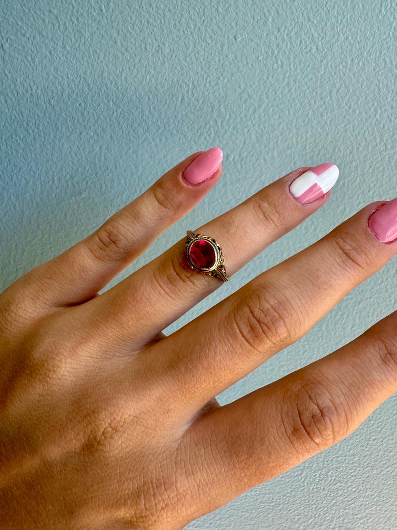 May include: A gold ring featuring an oval-shaped red gemstone set in a decorative metal frame. The band has intricate filigree detailing along the sides, surrounding the central stone which is held in place by a simple metal bezel setting.