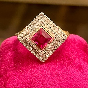 Könnte beinhalten: Ein quadratischer Ring mit einem großen, leuchtend pinkfarbenen Rubin, der von einem Heiligenschein aus kleineren Diamanten umgeben ist. Der Ring ist in eine goldene Fassung gefasst.
