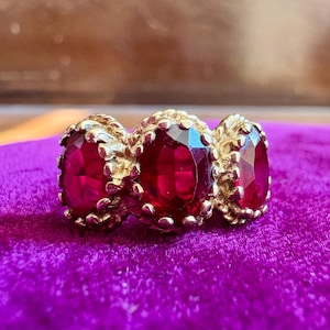 May include: A gold ring featuring three oval-cut deep red gemstones arranged horizontally. Each stone is secured in a prong setting with a textured, rope-like metal border surrounding the base of the gems, creating a continuous decorative band.