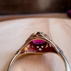 May include: A gold ring featuring an oval-cut vibrant pink gemstone set within an intricate, openwork metal gallery. The band is smooth and polished, while the setting displays detailed filigree patterns surrounding the central stone.