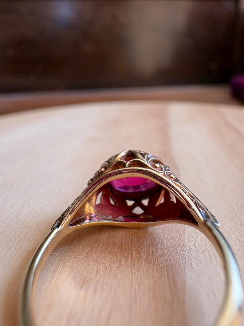 May include: A gold ring featuring an oval-cut vibrant pink gemstone set within an intricate, openwork metal gallery. The band is smooth and polished, while the setting displays detailed filigree patterns surrounding the central stone.