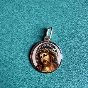 Może przedstawiać: Okrągła zawieszka z metalu w kolorze srebrnym, przedstawiająca kolorowy, szczegółowy portret Jezusa w koronie cierniowej. Obraz umieszczono na metalicznym tle z ozdobną, tłoczoną obwódką, całość posiada małe srebrne kółeczko do zawieszenia.