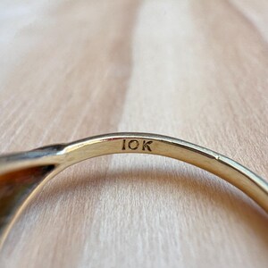 May include: A gold ring band featuring the engraved text 10K on the interior surface. The metal has a polished finish and a curved shape, showing the hallmark clearly stamped into the inner side of the band against a light wood grain texture.