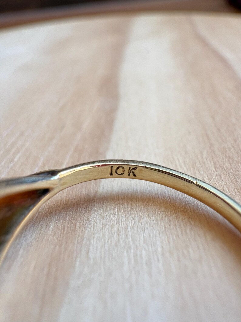 May include: A gold ring band featuring the engraved text 10K on the interior surface. The metal has a polished finish and a curved shape, showing the hallmark clearly stamped into the inner side of the band against a light wood grain texture.